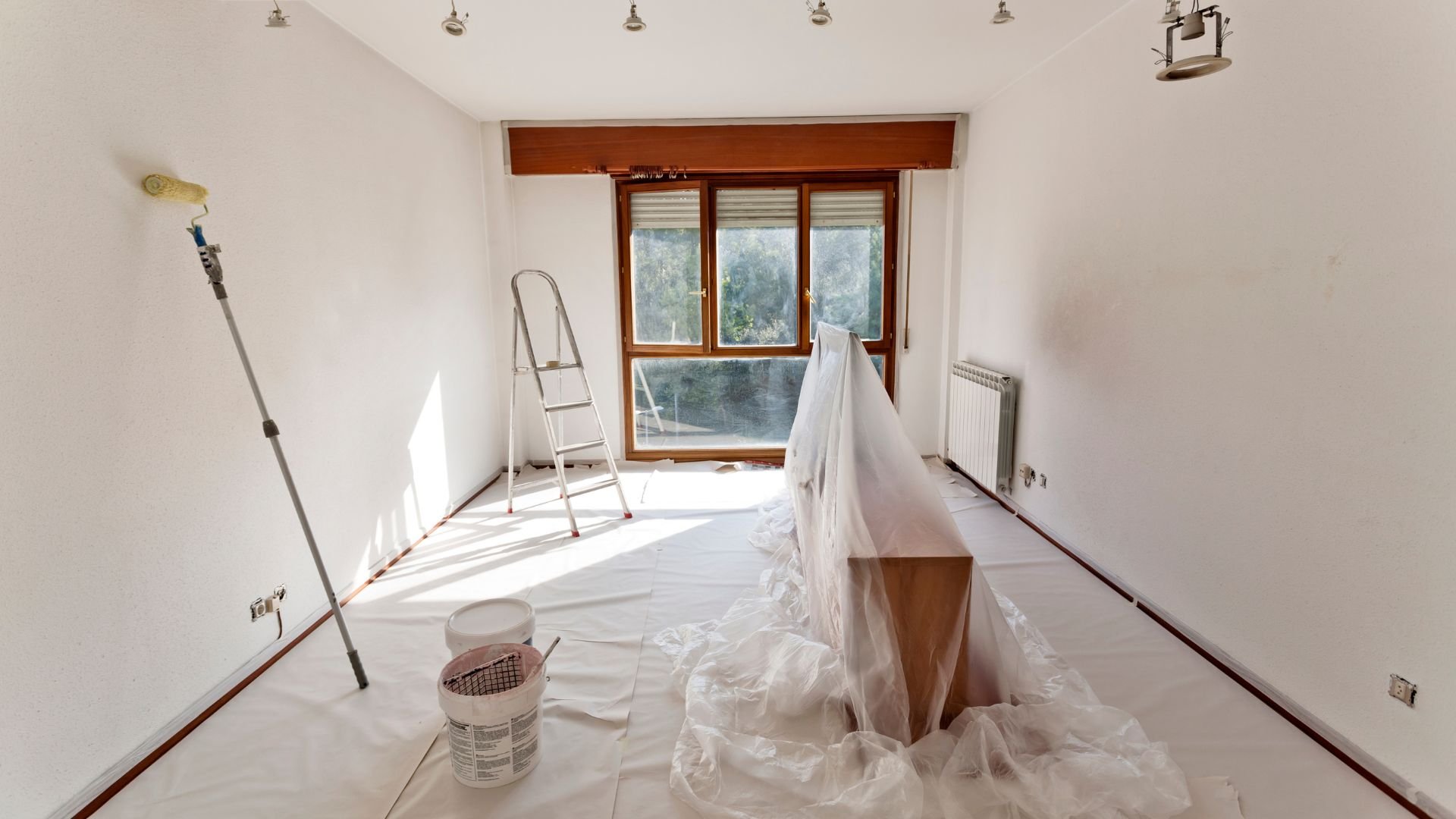 Room under renovation with paint supplies, ladder, and furniture covered in plastic