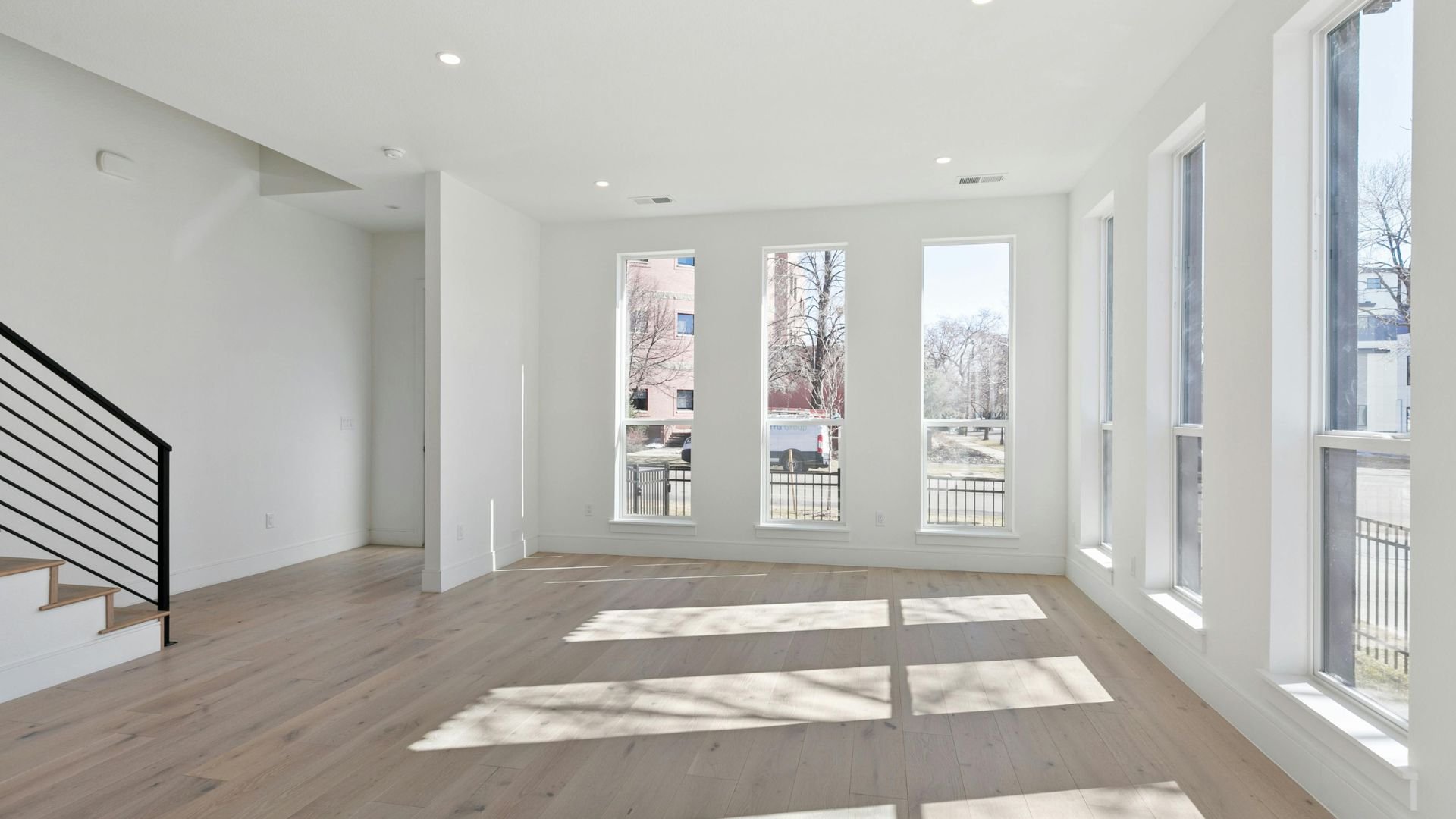 Empty bright room with large windows, wooden floor, and modern staircase